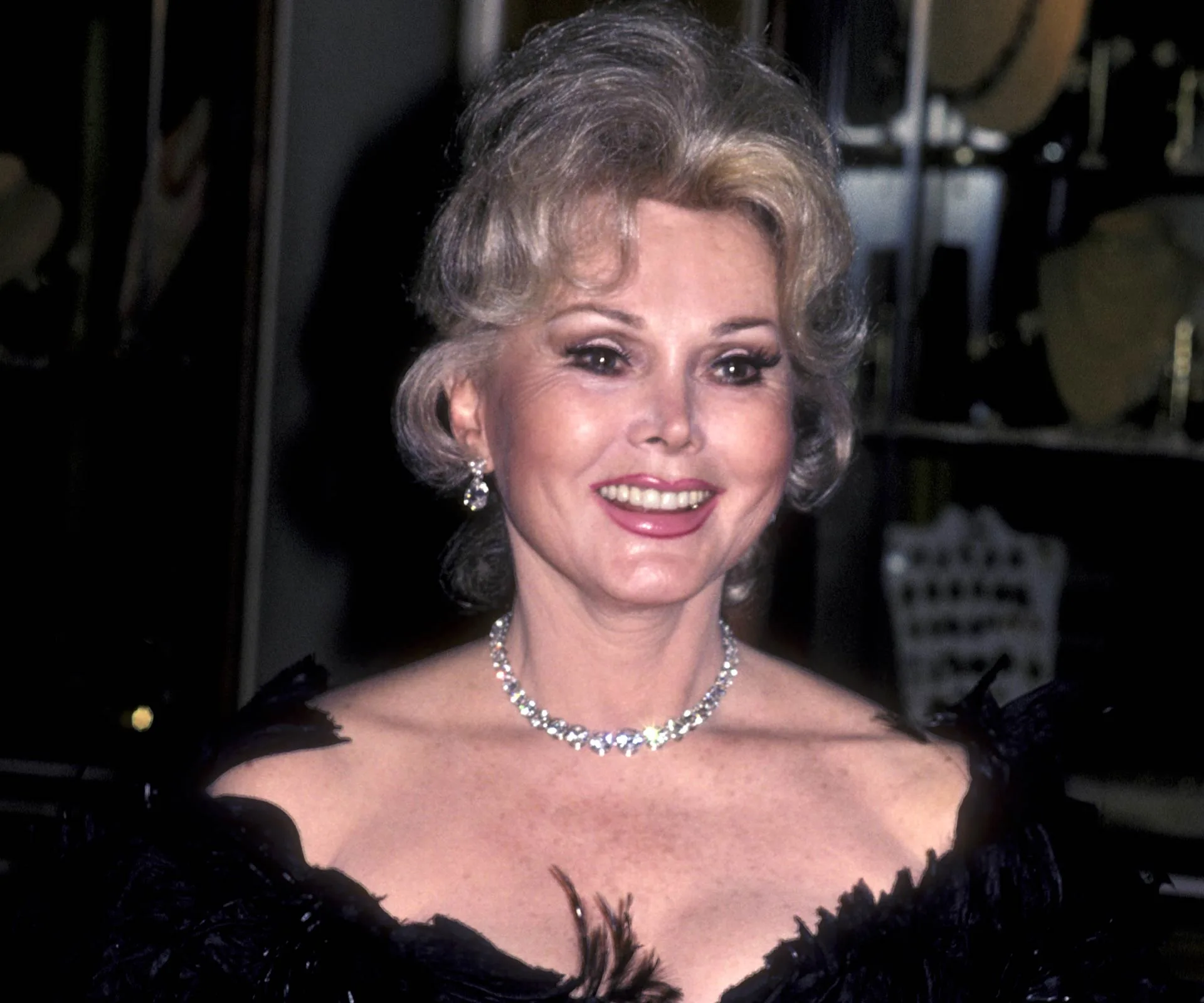 Zsa Zsa Gabor smiling, wearing a black dress and diamond necklace at an event.
