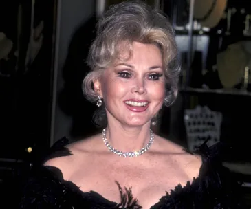 Zsa Zsa Gabor smiling, wearing a black dress and diamond necklace at an event.