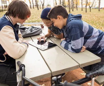 Three boys at a picnic table in a park, focused on a tablet screen.