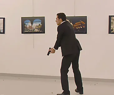Man in a suit holding a gun at an art gallery with framed pictures on the wall.