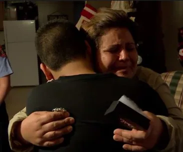A woman emotionally embraces a young boy who is facing away, in a warmly lit room.