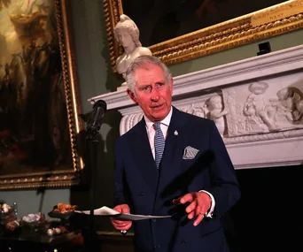 Prince Charles speaks at a lectern with a microphone, in front of ornate artwork and a decorative mantelpiece.