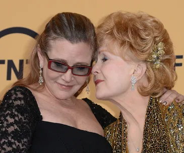 Two women embracing, with one wearing glasses and the other in a gold outfit, showing a close affectionate moment.