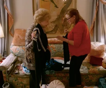 Two women in a cozy room, engaged in a lively conversation, each holding a piece of clothing.