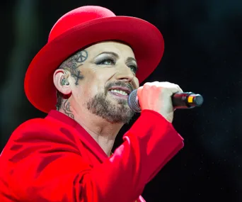 "Boy George performing on stage, wearing a red outfit and hat, holding a microphone."