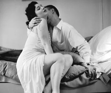 A couple embraces intimately on a bed, with the woman in a slip and the man in a shirt, in a black-and-white photograph.