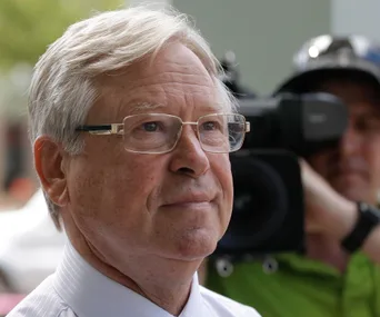 Elderly man with glasses, looking thoughtful, with a cameraman in the background.