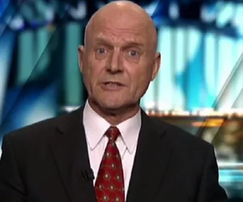 A man in a suit and red tie speaking on a news program with a blurred background.