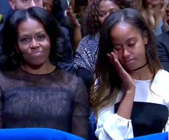 Michelle Obama and her daughter sit in an audience; Michelle smiles, while her daughter appears emotional and wipes her eye.