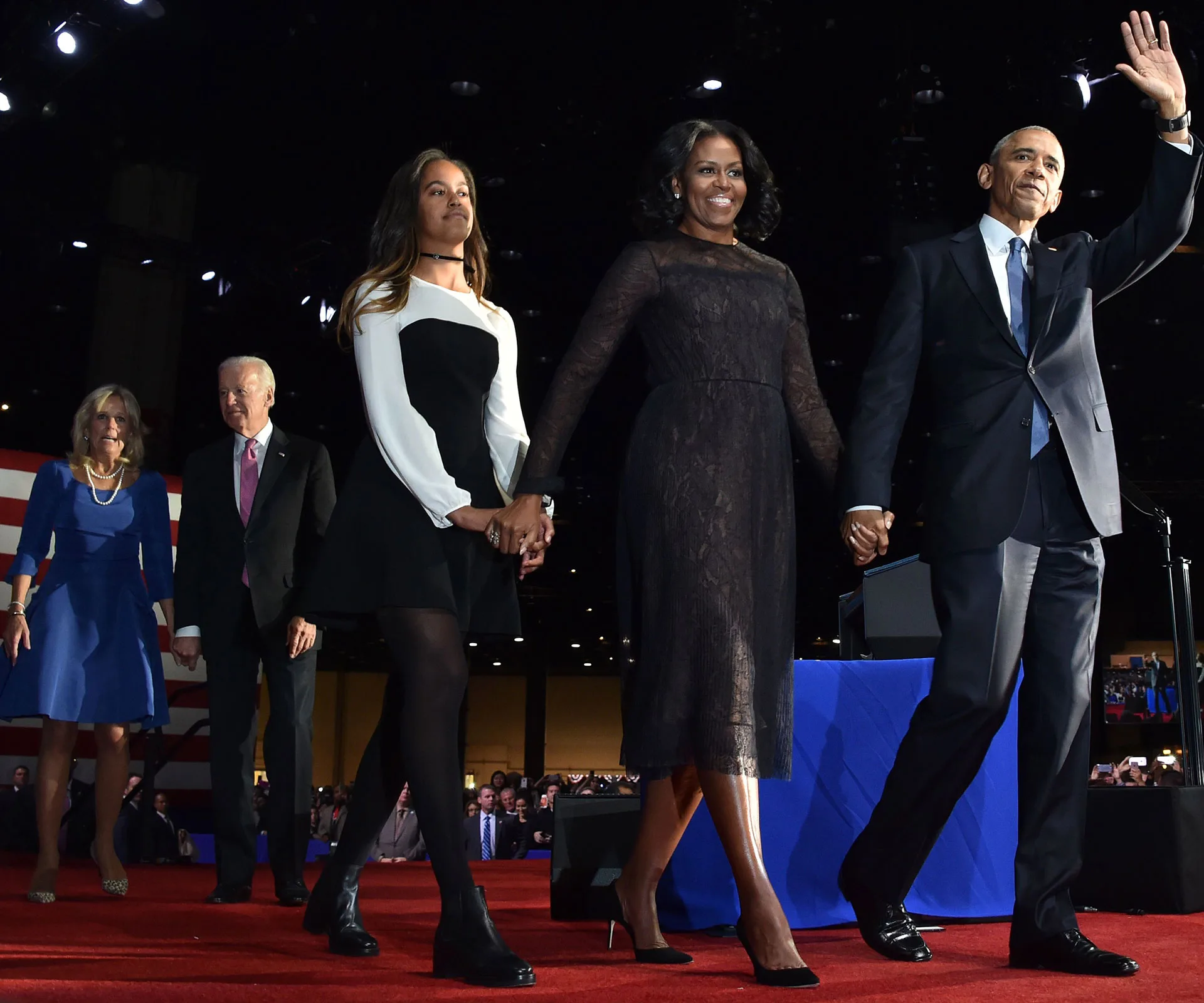 Former U.S. President and First Lady, with others, walk on stage holding hands at a public event.