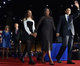 Former U.S. President and First Lady, with others, walk on stage holding hands at a public event.