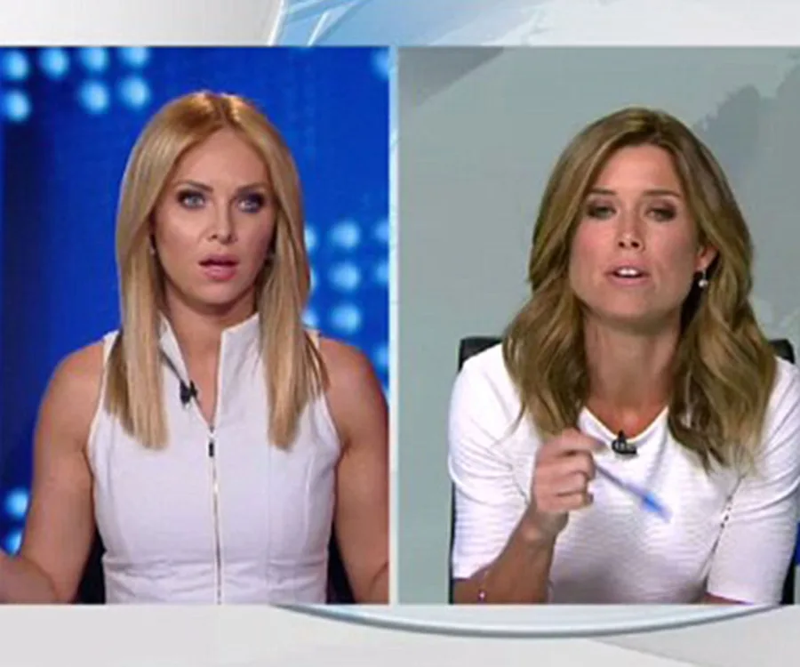 Two news presenters in white outfits during a live broadcast, one appears surprised, the other is gesturing with a pen.