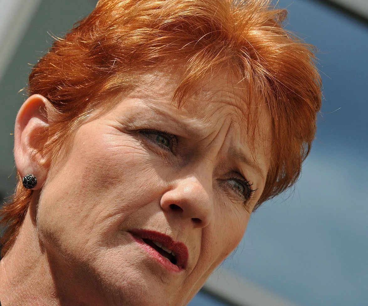 Red-haired person with a serious expression, captured in close-up against a blue sky background.