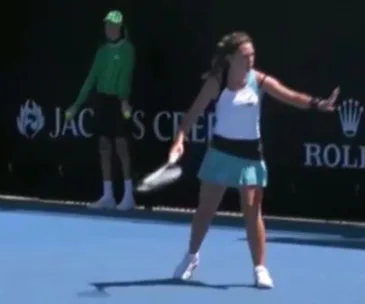 Tennis player gestures on court, ball boy stands in the background during a match.