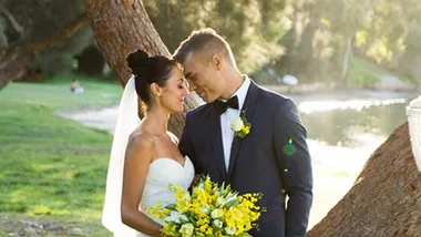 Bride and groom standing close by a tree, holding a yellow bouquet, with a sunny park background.