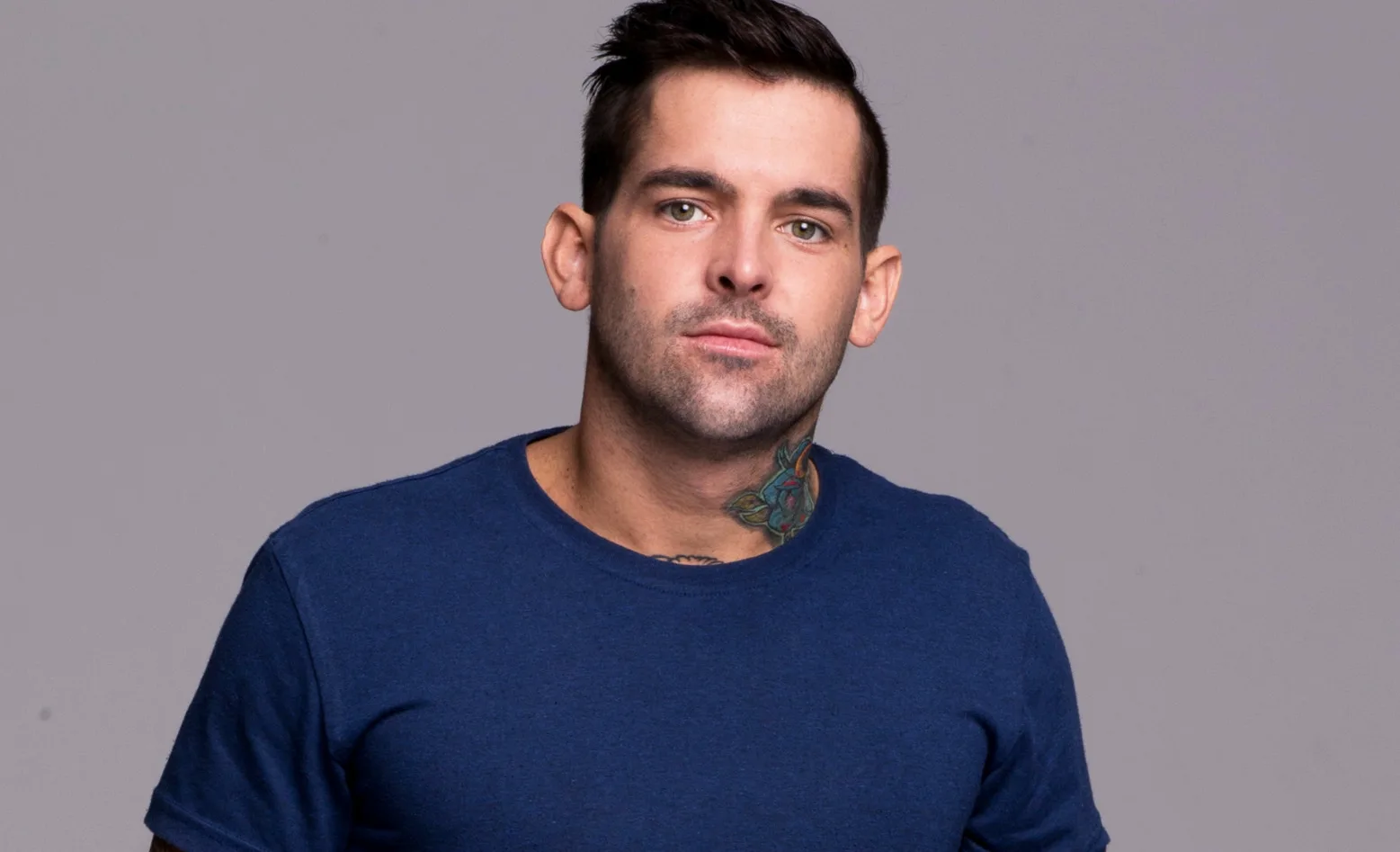 Man in a blue shirt with a colorful neck tattoo stands against a gray background.