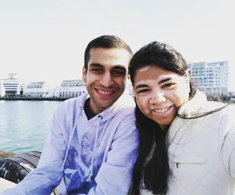 A smiling couple at a waterfront, the woman has vitiligo, with modern buildings visible in the background.