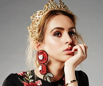 A person wearing a golden crown and large floral earrings, gazing thoughtfully with their hand on their chin.