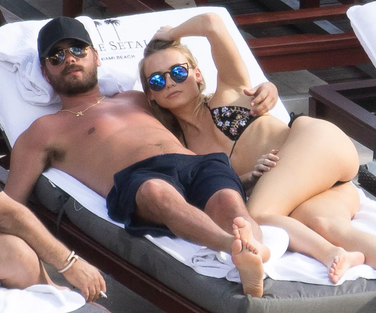 Two people lounging on a sunbed by a pool, wearing swimwear and sunglasses, appearing relaxed.
