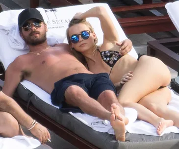 Two people lounging on a sunbed by a pool, wearing swimwear and sunglasses, appearing relaxed.