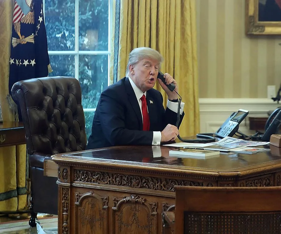 Man talking on phone at a large wooden desk in an office with yellow curtains and a window in the background.