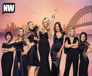 Seven women in elegant black dresses posed against a cityscape, with a bridge in the background, under a sunset sky.