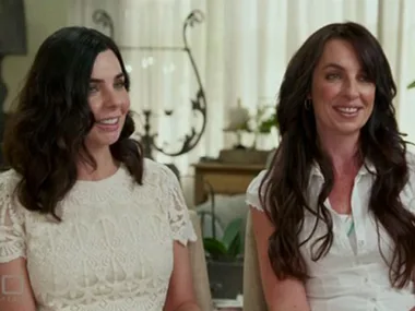 Two women with long dark hair smiling, sitting indoors, with decorative background elements.