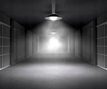 Dark hallway with barred prison cells on either side, dimmed overhead lights, and a bright light source at the far end.
