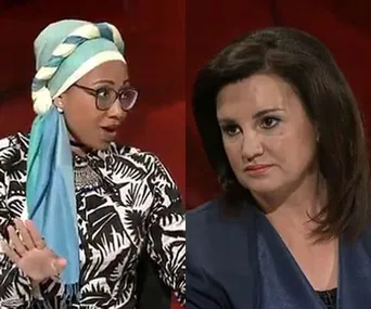 Two women in a discussion; one wearing a patterned headscarf, glasses, and print jacket; the other with dark hair and a blue top.