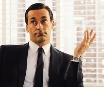 A man in a suit gestures with indifference, sitting in front of a window with blinds.