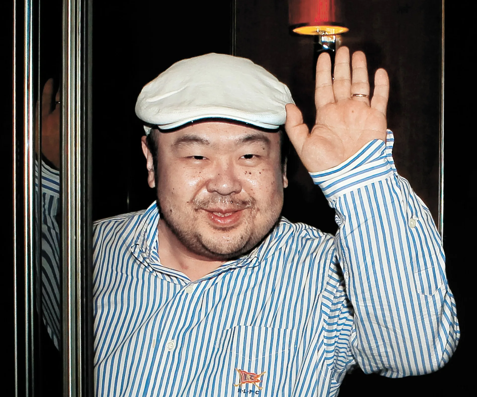 Man in a striped shirt and flat cap waves with a slight smile.
