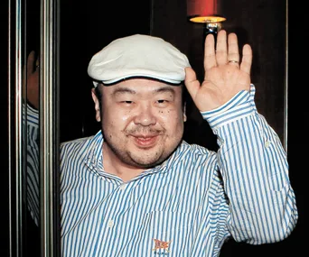 Man in a striped shirt and flat cap waves with a slight smile.