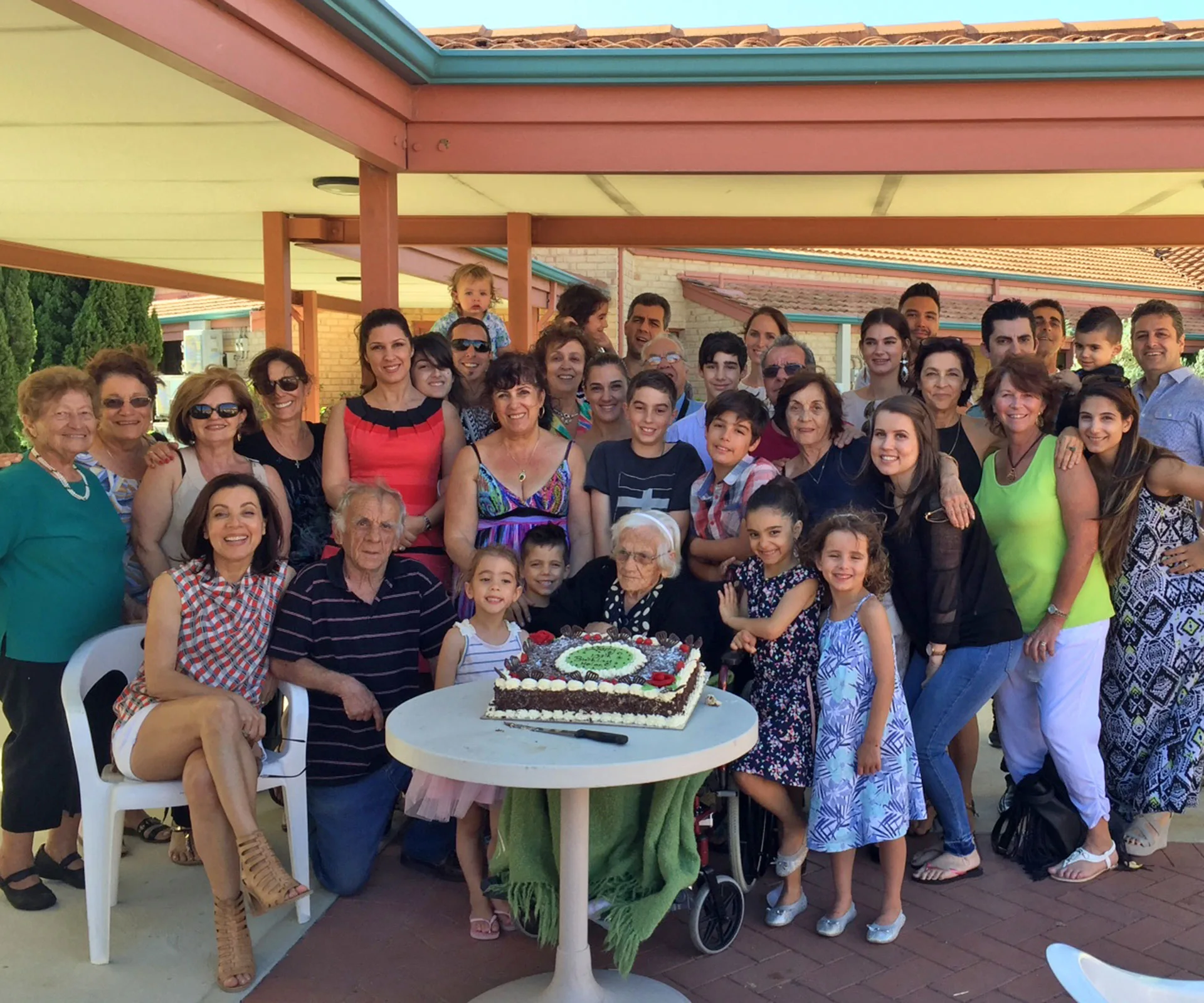 A large family gathers around a 110-year-old woman sitting in front of a cake at an outdoor celebration.