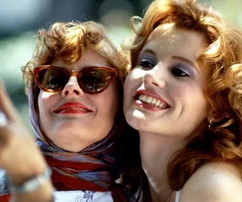 Two women smiling, one wearing sunglasses and a scarf, taking a selfie. Scene from the movie "Thelma & Louise".