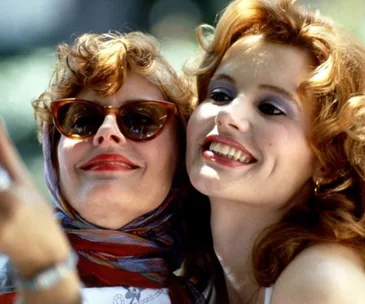 Two women smiling, one wearing sunglasses and a scarf, taking a selfie. Scene from the movie "Thelma & Louise".