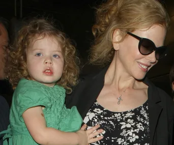 Woman in sunglasses smiling, holding a young child in a green dress with curly hair.
