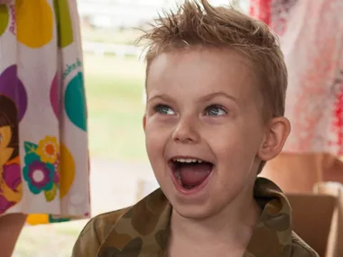 Young boy with spiky hair smiling widely at a party, wearing a camouflage jacket.