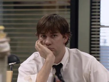 A man in an office setting, wearing a white shirt and black tie, rests his face on his hand, appearing bored.