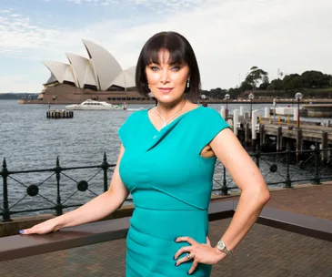 Woman in teal dress posing in front of Sydney Opera House with water and a pier in the background.