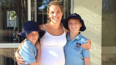 A smiling woman stands with two children in school uniforms and hats, outdoors near a building.
