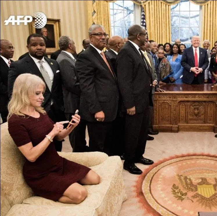 Kellyanne Conway on Oval Office couch during meeting with standing guests and man near Resolute Desk.