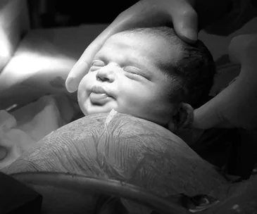 Newborn baby being held gently, freshly delivered, with eyes closed and tongue out, under focused lighting.