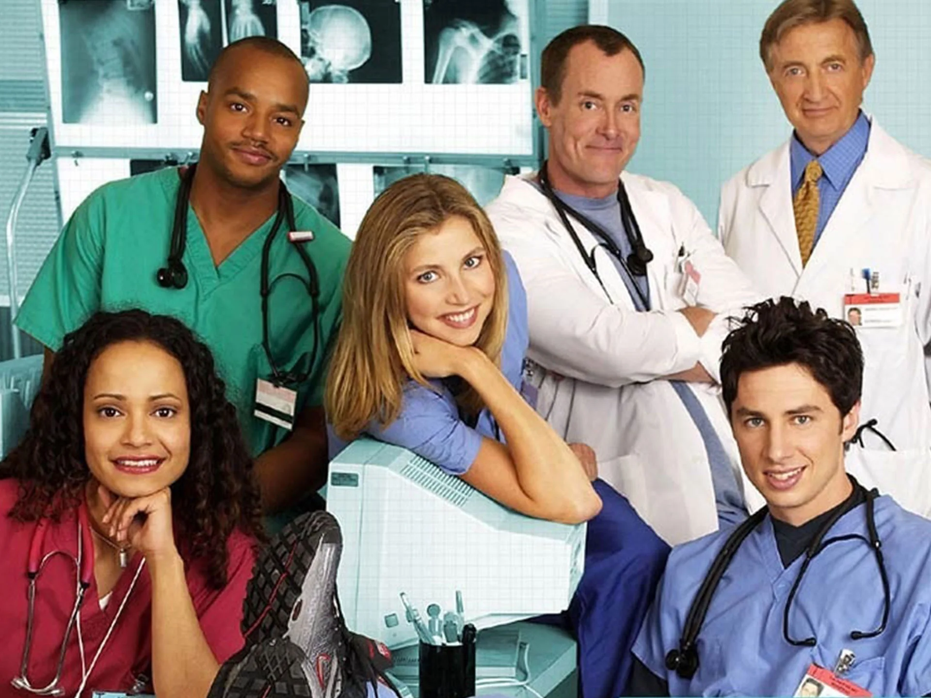 Cast of "Scrubs" in character, smiling in a hospital setting with medical equipment and x-rays in the background.