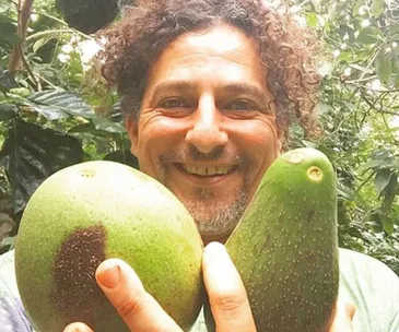 A smiling person holding two large avocados in a lush garden setting.