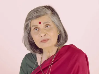 Elderly woman with gray hair in a saree, wearing a bindi, against a plain background.