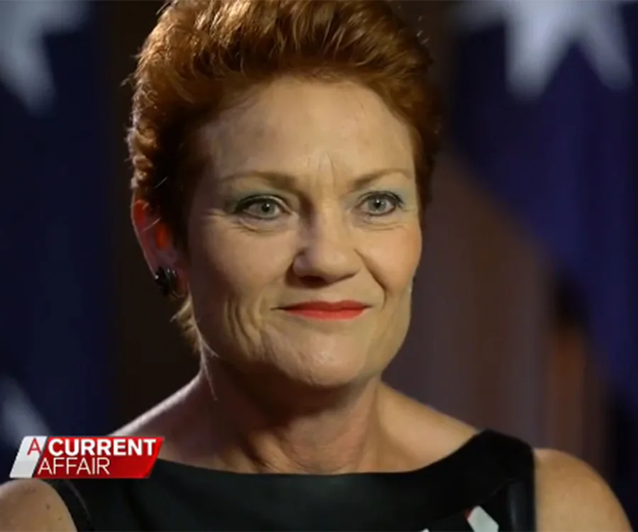Close-up of a woman with short reddish hair, speaking on a TV program with flags in the background.