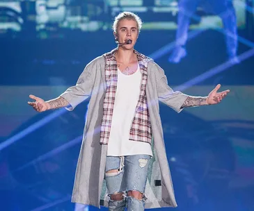 A performer with a microphone, wearing a plaid shirt and ripped jeans, extends arms on stage during a concert.