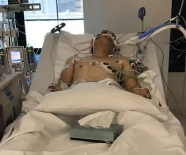 A person with medical equipment is lying in a hospital bed, connected to various monitors and tubes.
