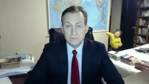 Man in suit on video call; child in yellow enters room playfully from the background.