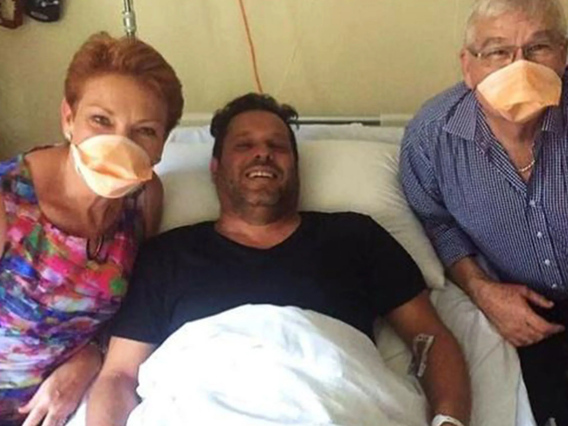 Three people in a hospital room, two wearing masks, visiting a patient lying in bed, smiling.
